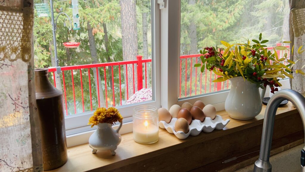 Affordable fall flower bouquet on a kitchen windowsill made from wild foliage clippings and wild rose hips in a white pitcher next to fresh eggs, a lit candle and a tiny creamer pitcher of orange chrysanthemums.