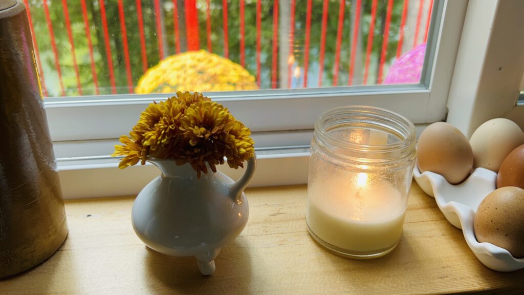 Mini bouquet of orange chrysanthemums in a tiny white pitcher next to a lit candle on kitchen windowsill.