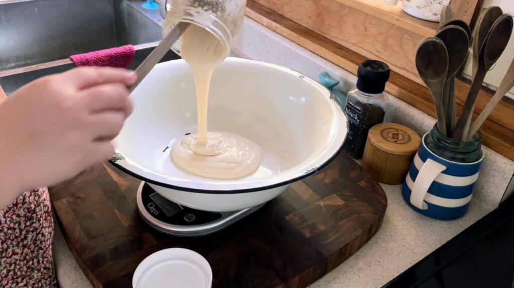 Pouring sourdough starter from a mason jar into a big white enamelware bowl on a kitchen scale.