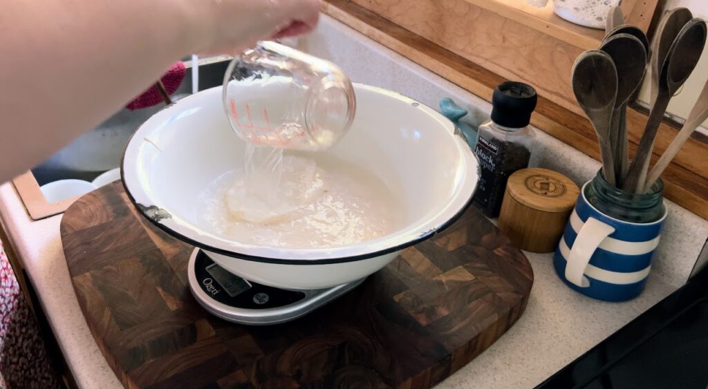 Pouring water from a glass measuring cup into a large white enamelware bowl with sourdough starter. 