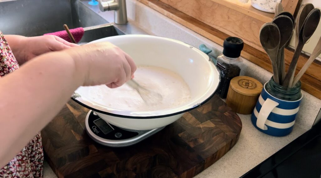 Whisking water and sourdough starter together in a large white enamelware bowl.