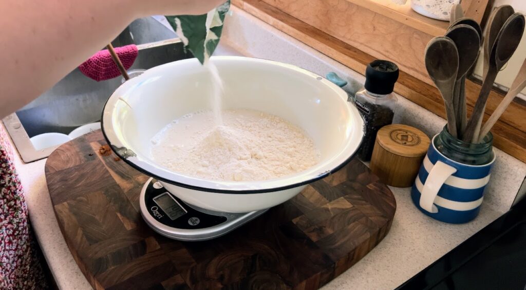 Measuring all-purpose flower into sourdough starter and water in large white enamelware bowl.