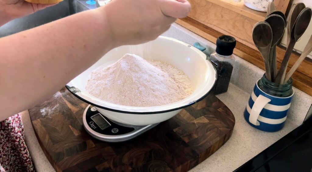 Measuring salt into large white enamelware bowl with flour and sourdough starter.