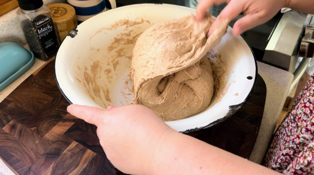 Stretching and folding whole wheat sourdough dough in a large white enamelware bowl.