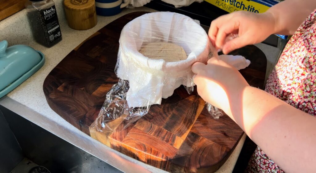 Putting plastic wrap over a bowl with a white kitchen towel and sourdough dough for proofing. 