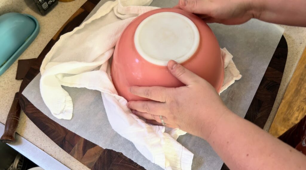 Pink vintage pyrex bowl upside down with white kitchen towel on a wood cutting board.