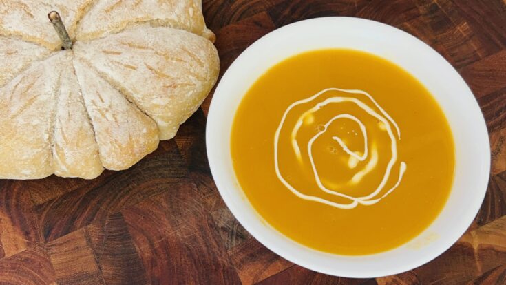 Bowl of roasted butternut squash soup in a white bowl with a drizzle of heavy cream next to a pumpkin shaped loaf of sourdough bread.