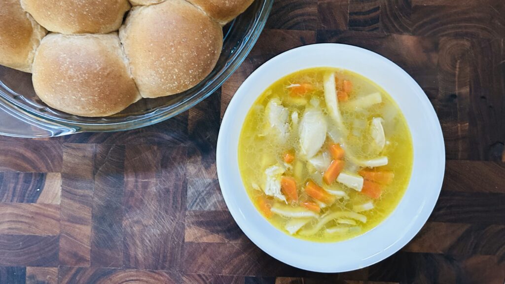 Bowl of chicken noodle soup with egg noodles, chicken, carrots, onion and celery, in a white bowl next to a round glass dish with honey whole wheat dinner rolls. 