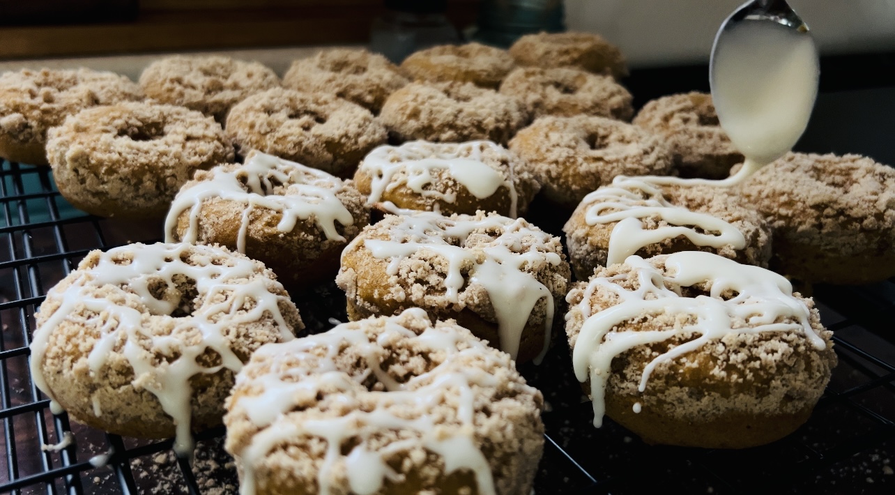 Baked sourdough pumpkin donuts topped with a streusel crumb topping and being drizzled with a simple powdered sugar frosting.