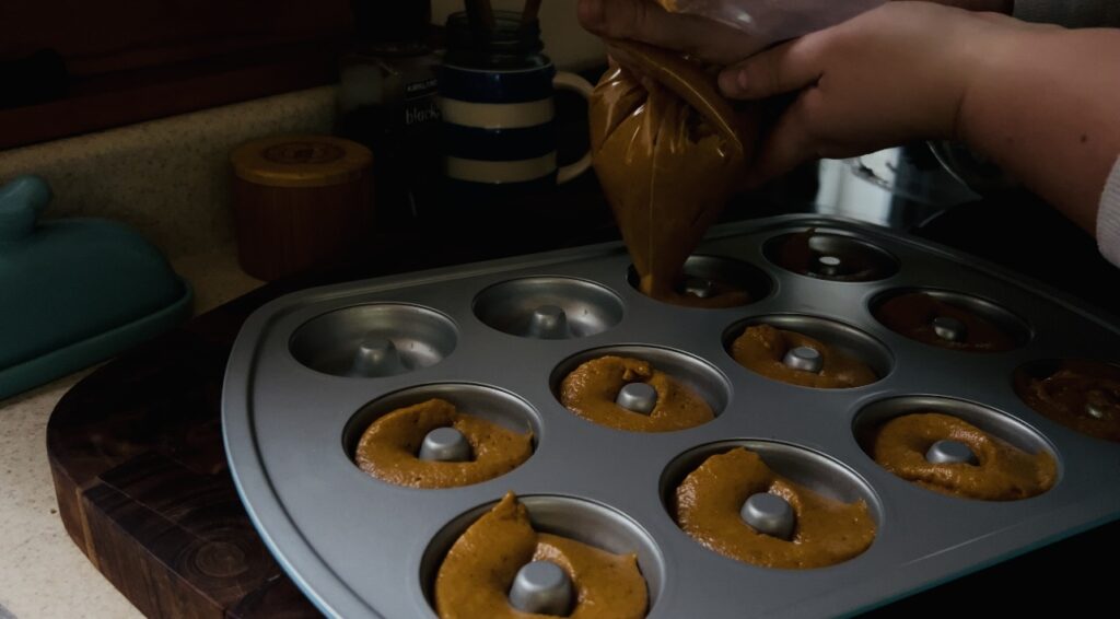 Piping sourdough pumpkin donut batter into a donut pan with a large ziploc bag used as a pastry bag.