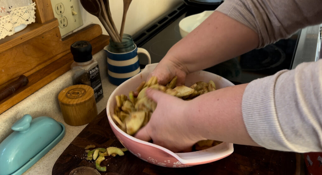 Mixing a sugar mixture and chopped granny smith apples together in a large pink pyrex bowl for rustic apple pie.