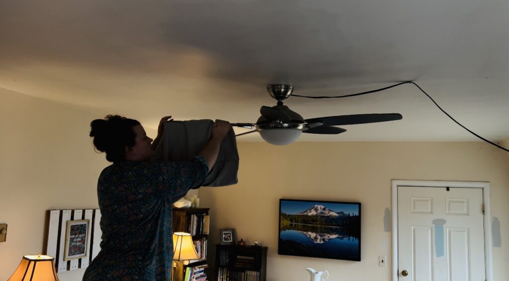 Woman using a pillow case to dust off the blades of a ceiling fan.