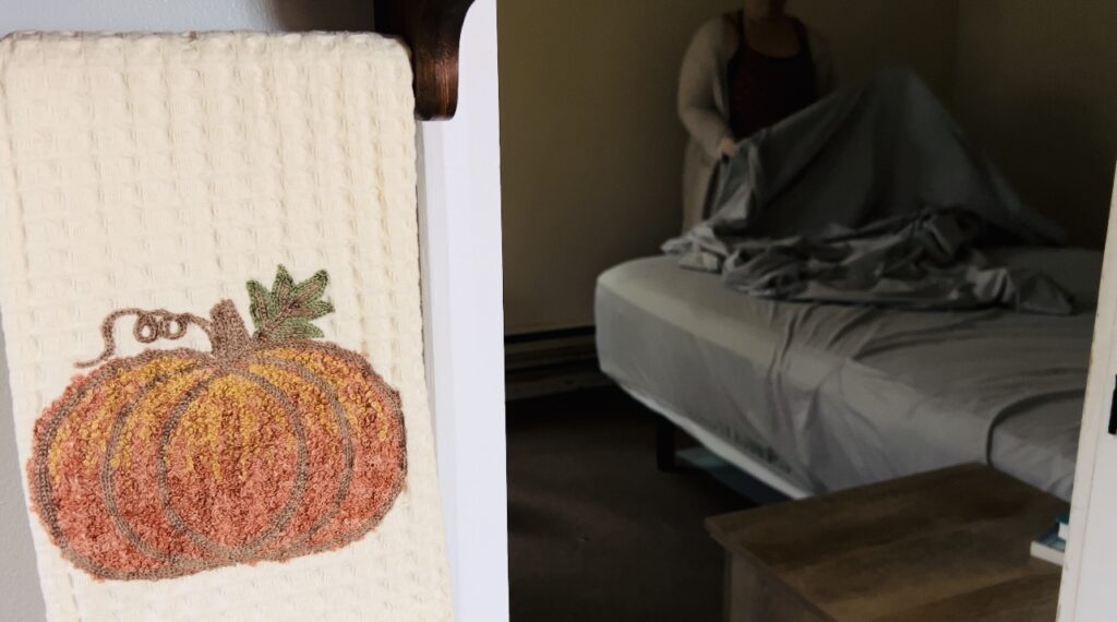 Woman in the background changing the sheets on her bed with a kitchen towel with pumpkin on it in the foreground.