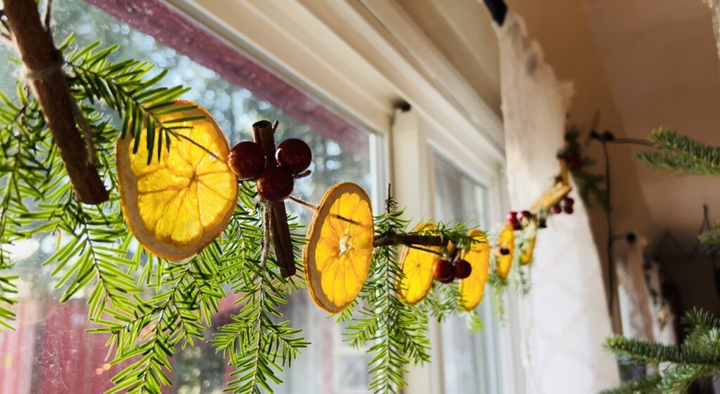 Handmade garland made of dehydrated lemon slices, cinnamon sticks, hemlock sprigs and fake red berries strung onto brown twine and strung across a window.