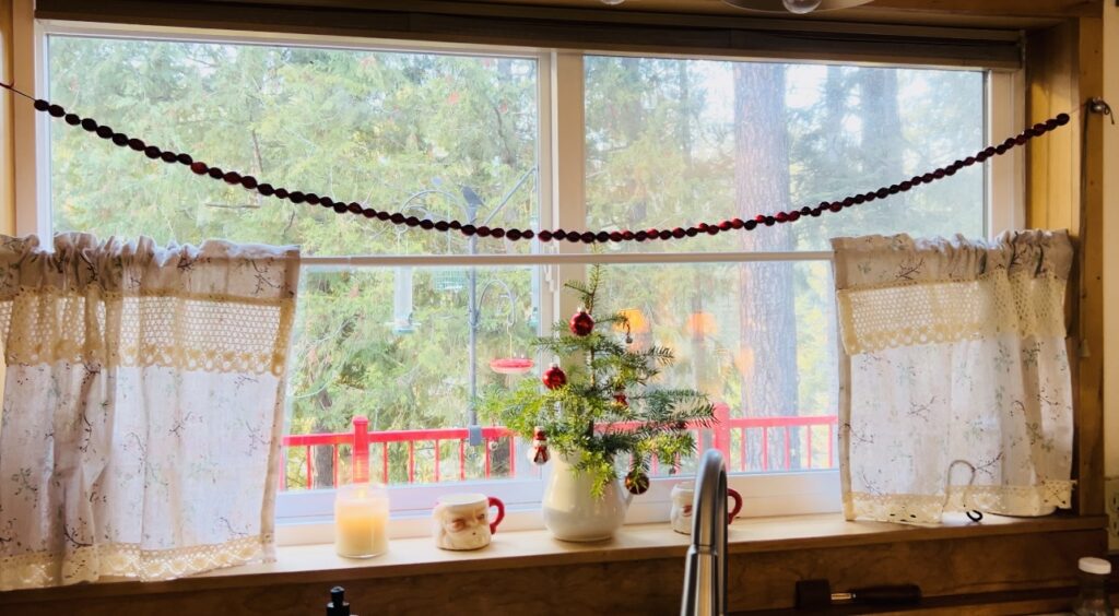 Kitchen window decorated for Christmas with a small Christmas tree, Santa mugs, a candle and a string of fresh cranberries.