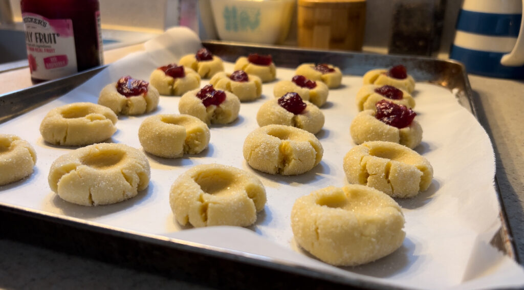 Cookie sheet with raw thumbprint cookies getting their raspberry jam filling. 