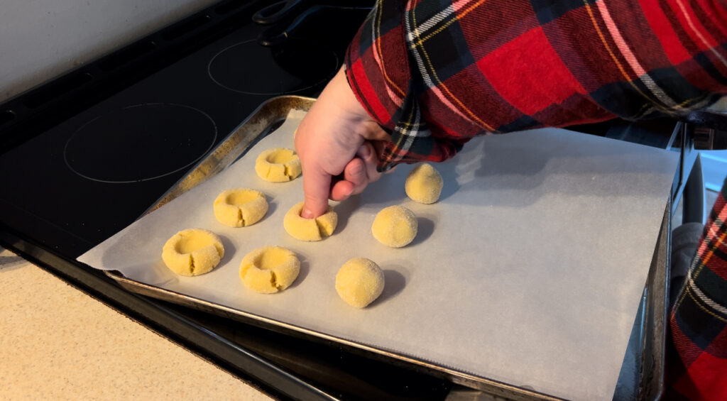 Woman wearing red and green plaid shirt pressing her thumb into cookie dough to make thumbprint cookies.