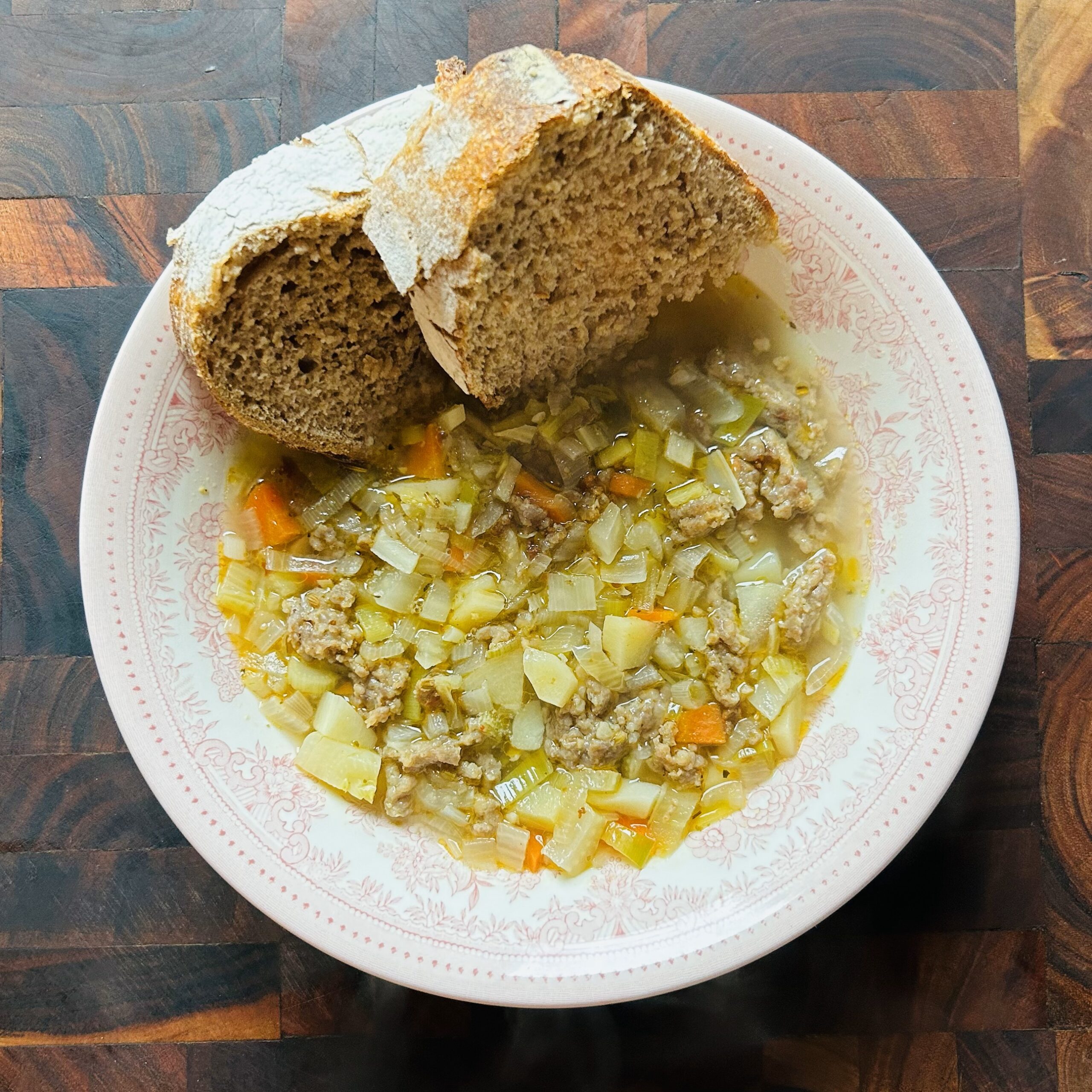 Bowl on winter vegetable and sausage soup in a white bowl with a slice of hearty whole wheat sourdough bread.