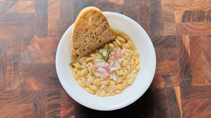 Bowl of white bean and shallot soup with rosemary and garlic with a slice of whole wheat sourdough bread.