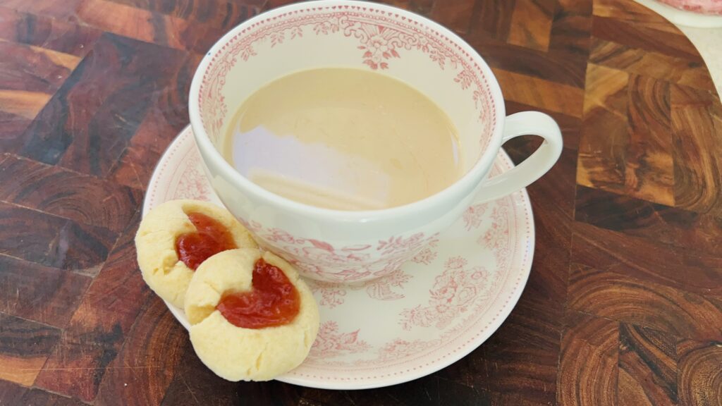 Two strawberry thumbprint cookies on a saucer next to a cup of tea.