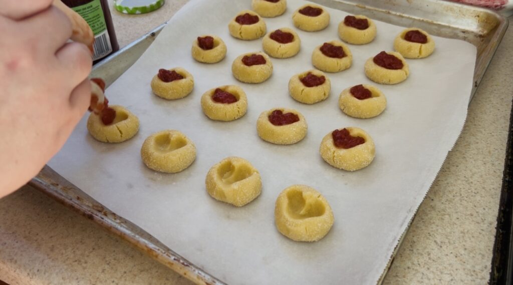 Placing strawberry jam into the indentation in strawberry thumbprint cookies before baking.