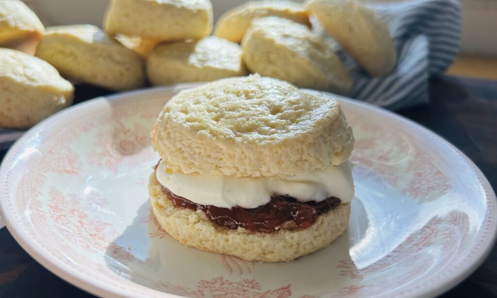 Freshly baked classic scone with strawberry jam and easy mock clotted cream on a plate with scones piled in the background. 