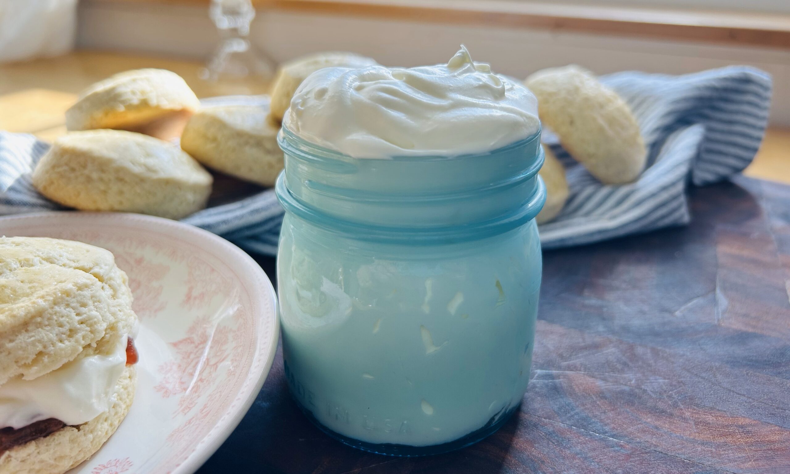 Easy mock clotted cream in a small blue mason jar with a freshly baked scone with strawberry jam and clotted cream on a plate next to it with more scones in the background.