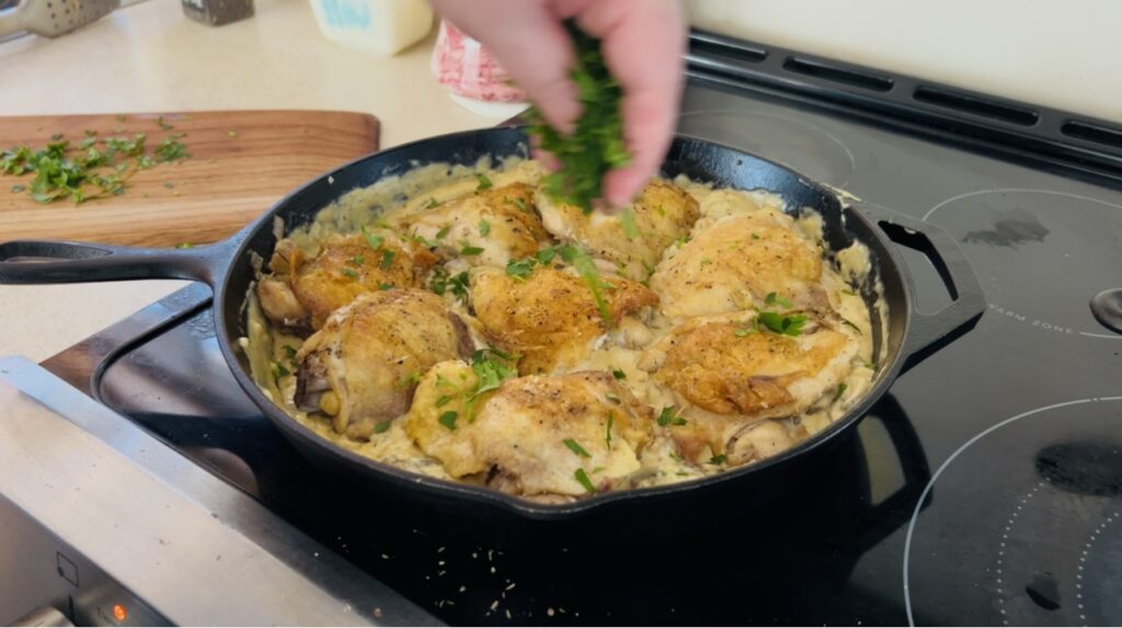 Chicken thighs in cream sauce being topped with freshly chopped parsley. 