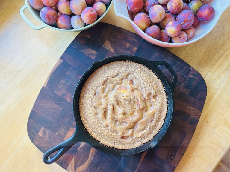 Freshly baked plum cobbler made in a cast iron skillet with bowls of plums around it.